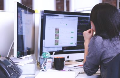 A female office worker is looking at two display monitors at her work desk.