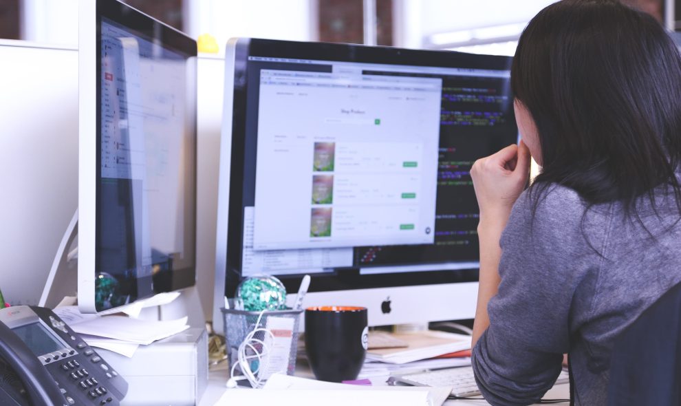 A female office worker is looking at two display monitors at her work desk.