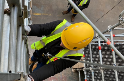 A warehouse worker wearing a hard hat and safety vest using a ladder at work.