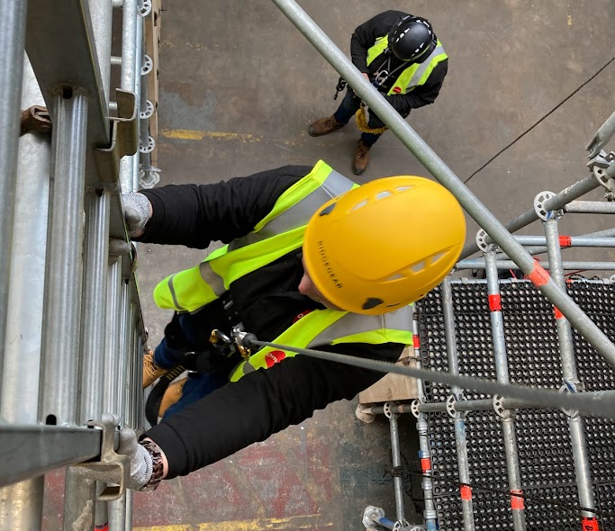 A warehouse worker wearing a hard hat and safety vest using a ladder at work.